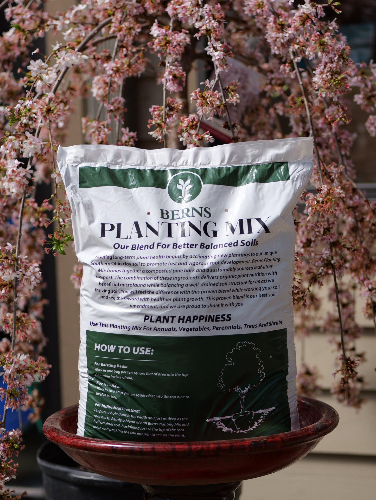 A bag of Berns Planting Mix soil sitting on a pot next to a potted plant with pink flowers.